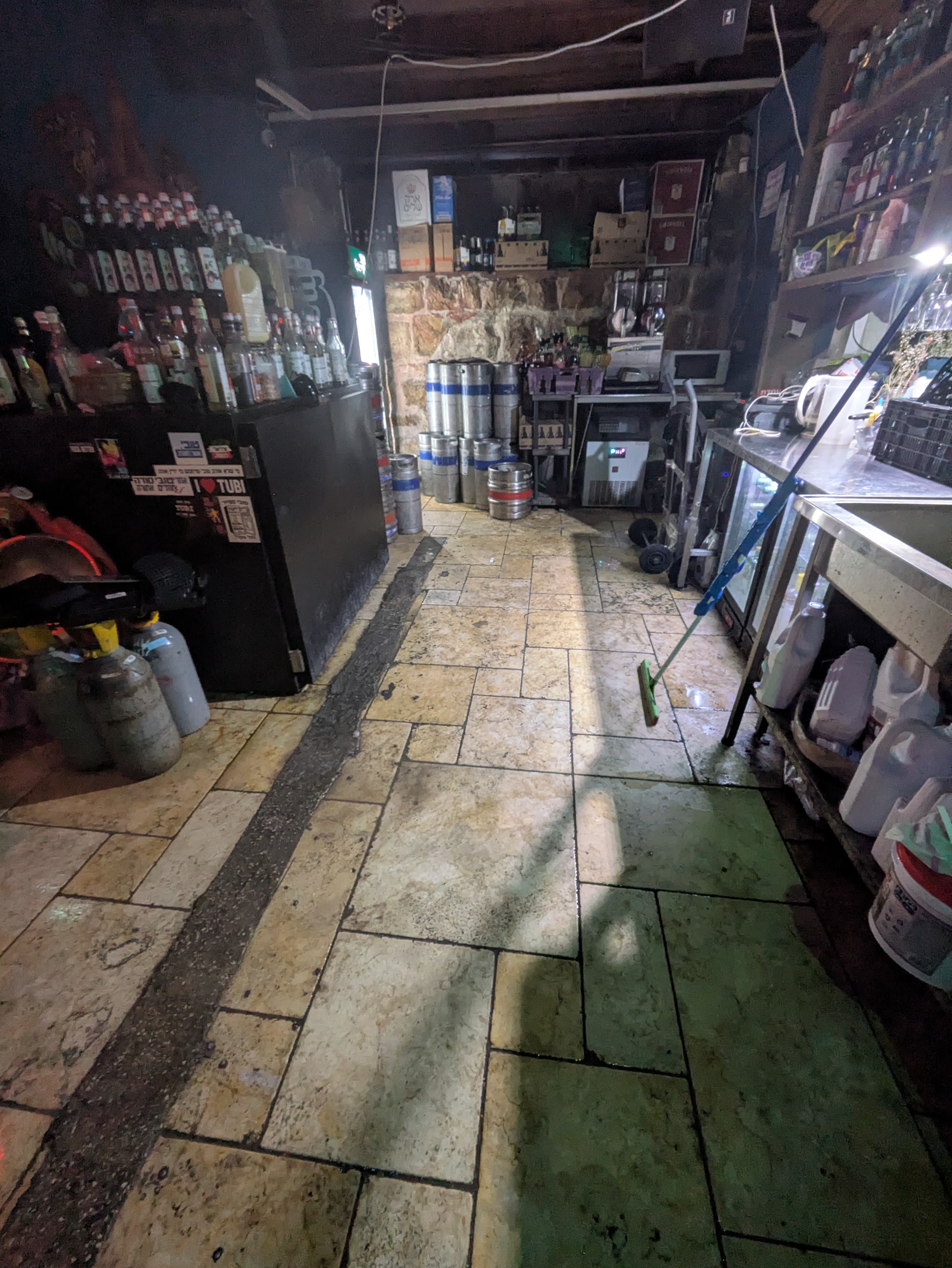 Dimly lit bar storage area with stone floors, metal kegs, and many liquor bottles.
